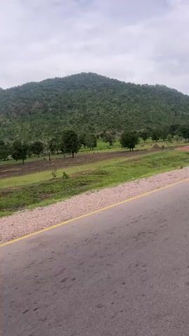 This is a video captured from a moving vehicle on a paved road in Minda, Nigeria. The foreground is dominated by the asphalt road, marked with a yellow line on the right edge. Beyond the road is a strip of grassy verge, followed by cultivated fields and scattered trees. In the background, a large, lush green hill rises prominently, covered in dense vegetation. The sky is overcast, suggesting a cloudy or possibly rainy day. The overall scene depicts a rural landscape with a focus on the natural environment and agricultural activity. There are no visible people or specific objects that stand out, beyond the general elements of a road, fields, trees, and a hill. The time of day appears to be daytime, given the natural light. The emotions conveyed are neutral, offering a glimpse of the scenery. There is no visible text in the video.