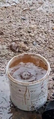 The video shows a dirty, white bucket filled with swirling brown water. The bucket has some stains and markings on its exterior. It sits on a muddy, uneven surface with scattered rocks and debris. There are sounds of running water in the background.