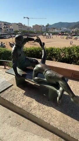 The video opens with a close-up of a bronze sculpture of two figures sitting on a bench. The sculpture is positioned in the foreground, with a beach and town in the background. The camera then pans to the right, revealing a busy beach scene under a clear blue sky.

The beach is filled with people sunbathing, relaxing under colorful umbrellas, and walking along the shore. The sand is golden, and the sea is calm and blue. In the distance, buildings of a town ascend a hillside, with lush green vegetation covering the slopes. A construction crane is visible to the left of the town.

The overall atmosphere is one of a bright, sunny day at a popular tourist destination. The scene is lively and vibrant, suggesting a vacation or holiday setting. The time of day appears to be late morning or early afternoon, given the strong sunlight and long shadows cast by the sculpture. The location is identified as Tossa de Mar, Spain, a coastal town known for its beaches and historic charm.

The sculpture itself depicts two individuals, one appearing older, with the other younger, engaged in conversation or sharing a moment. The bronze material and detailed craftsmanship suggest it is a significant piece of public art. The juxtaposition of the static sculpture with the dynamic human activity on the beach creates an interesting contrast.

No visible text is present in the video. The focus is on the visual experience of the location and the sculpture. The weather is clearly sunny and warm, ideal for a beach day. The emotions conveyed are those of relaxation, enjoyment, and perhaps a sense of historical connection suggested by the sculpture.