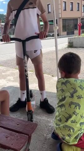 A young boy stands on a scooter, looking down at it. He is wearing a tan and white t-shirt, tan shorts, white socks, and black shoes. He has a black crossbody bag slung over his shoulder. Another young boy sits on a bench to the left, wearing gray shorts and a tan t-shirt. To the right, another boy wearing a neon green tank top with a zebra print and red pants is also sitting. The first boy grips the handlebars of the scooter and bends forward, as if preparing to ride. The background shows buildings, trees, and a street. The lighting suggests it is daytime.