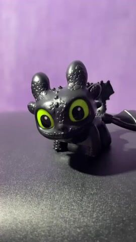 A black figurine of Toothless from How to Train Your Dragon is positioned on a dark, textured surface. The figurine has large, bright green eyes and detailed scaly skin. The background is a solid purple color. The camera is positioned slightly above and to the side of the figurine, and the lighting illuminates the subject clearly. The video shows subtle movements and slight shifts in focus, suggesting the camera or figurine may be gently nudged or the lighting is being adjusted.