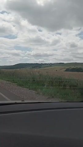 The video shows a view from inside a vehicle looking out at a rural landscape. A paved road is visible in the foreground, with a gravel shoulder. Beyond the road is a field of dry grass and some green vegetation. In the distance, there are rolling hills with some scattered buildings and trees. The sky is cloudy with patches of blue sky visible. A white sign with a red triangle and a black arrow pointing to the right is visible on the left side of the road. The video appears to be shot while the vehicle is moving.