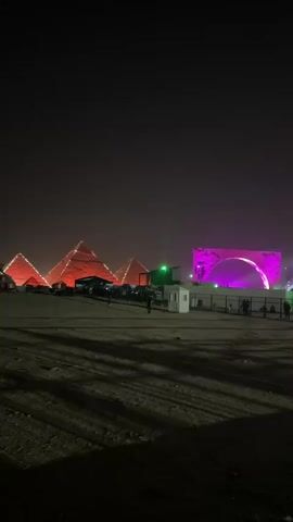 At night, the three lit-up pyramids are visible in the background. They are lit up with a red light. A large light-up arch is visible on the right side of the frame, with spotlights shining out of it. The foreground is a dirt road, with shadows created from lights. Some people are walking and standing in the foreground. There is a white, small security booth near the right side of the frame. The sound of music can be heard in the video.