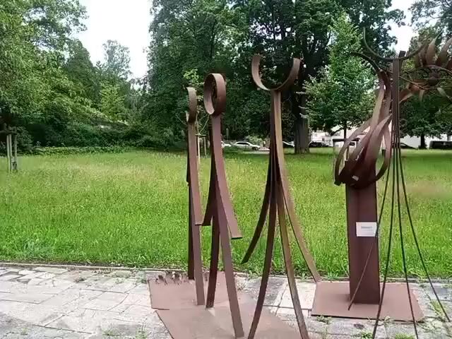 A video pans across a collection of abstract metal sculptures set on a stone pathway in front of a grassy field with trees. The sculptures are made of rusted metal and vary in shape and size, with some resembling figures and others abstract forms. The environment appears to be an outdoor park or public space.