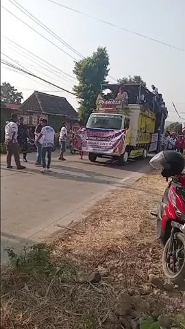 A yellow truck with a raised platform is driving down a paved road. People are standing on the platform of the truck and on the side of the road. There are banners on the front of the truck with text in Indonesian. A red motorcycle is parked on the side of the road in the foreground. The sky is clear and the sun is shining. The area appears to be a village or rural setting with houses and trees visible in the background.