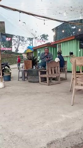 The scene unfolds under a white canopy strung with bare-bulb lights, illuminating an outdoor sitting area. In the foreground, a weathered wooden chair stands slightly apart. Moving towards the center, two more wooden chairs are positioned facing the camera, one occupied by a man in a blue plaid shirt and cap who is either smoking or speaking into a small device held to his mouth. A second man sits on the other chair, turned away from the camera. In front of them, several tires stacked atop one another serve as a makeshift table.

Behind them is a green structure, possibly a storage container repurposed as a small shop or kiosk. To the left of the scene, a motorbike is parked, partially obscured by plants in a blue bucket. A person in a red jacket walks away into the distance. The ground is bare concrete. Small Indonesian flags are strung up to the side. The backdrop is a view of trees.