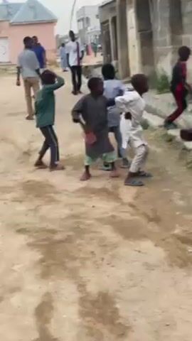 The video shows four young children in a residential neighborhood. They appear to be dancing to music coming from an unseen source. One child wears a white patterned outfit, another a gray outfit, and the third a green outfit with black pants. The fourth is partially hidden but appears to be wearing a darker outfit. Other people are walking in the background, including a man in a white and black jersey. The setting is a sandy street between buildings, with overcast lighting.