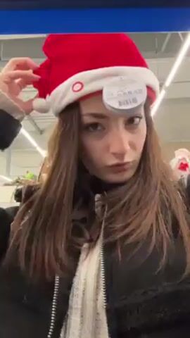 A woman with long brown hair, wearing a white sweater and a black jacket, and a red and white Santa hat, stands in an aisle of a store with shelves filled with Christmas decorations, including gingerbread houses.  She looks at the camera, pouts her lips, makes a peace sign, and smiles. Another person is visible in the background to her left.  The overhead lighting is bright and industrial. The camera is moving slightly, as if hand held.