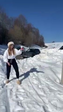 The video depicts a young woman throwing a snowball in a snow-covered environment under a clear blue sky. The woman is dressed in black leggings, a white hoodie, and a furry hat. She throws the snowball with her right hand and then immediately bends over, laughing, presumably because she missed her target. Behind her are several parked cars, large piles of snow, and bare trees. To her right is a gray utility pole. The snow is marked with footprints and the shadows of the trees and pole.