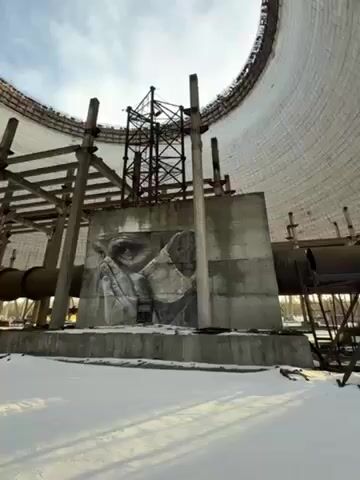 The camera slowly pans upward within a large, cylindrical concrete structure. Snow covers the ground. There is a large mural depicting an old woman with her hands clasped in prayer, painted on a concrete structure at eye level. A complex of metal beams and scaffolding extends upward, partially obscuring the view of the open top of the cylinder. The sky is visible through the opening, which is partially obscured by clouds.  Large pipes run horizontally along the lower part of the structure.