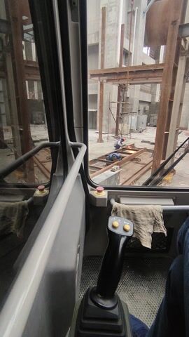 The video shows the view from inside the cab of a crane. Through the windshield, a construction site is visible.  The site contains a lot of metal beams and concrete structures. A worker is seen welding on a platform made of beams. The crane operator's controls are visible on the right of the screen.