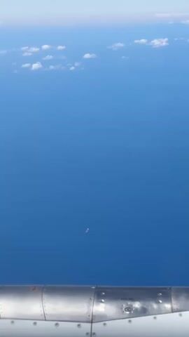 This is a video taken from an airplane window, looking down at a vast expanse of blue ocean. In the distance, a single small ship is visible on the water. The lower part of the video shows a portion of the airplane's engine, which is metallic and appears to be a turbine. The sky above is a pale blue with scattered white clouds. The video is brief, showing the unchanging scene of the ocean and the ship as the plane presumably flies over it.