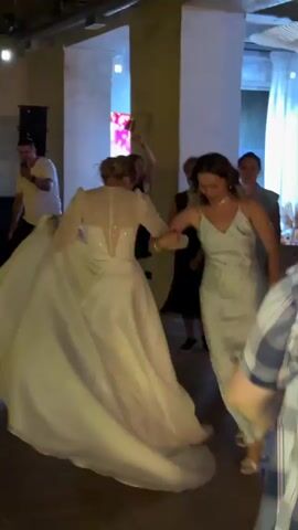 A wedding reception is underway, with a bride in a white gown dancing with guests. The bride is twirling, her long train trailing behind her. Several women are visible in the background, some of whom appear to be dancing or watching. The room is decorated with warm lighting, and a large pillar is visible on the left side of the frame. The floor is dark, and there are hints of red and blue lighting, suggesting a festive atmosphere.