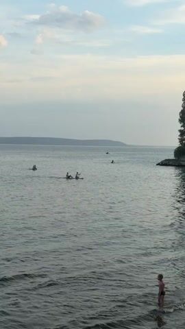 A video shows a wide shot of a body of water with several people engaged in various activities. In the foreground, a young boy stands at the edge of the water, facing away from the camera. The water is calm with a gentle ripple effect. In the mid-ground, two people are on a paddleboard, one paddling. Further out, another person is on a paddleboard. In the background, there are several other individuals on the water, some appearing to be on smaller watercraft. A distant shoreline with hills is visible under a hazy sky with scattered clouds. On the right side of the frame, there is a dense tree line and a rocky embankment leading into the water. The overall scene suggests a relaxed, outdoor environment.
