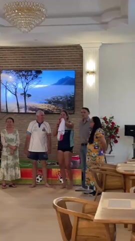 The video shows a group of five people standing in a room. A large television screen is mounted on a brick wall behind them, displaying a landscape image. In front of the television, a woman is holding up a white t-shirt. She is wearing a dark blue and white athletic top and dark shorts. To her left, a man in a white polo shirt and blue shorts stands with his hands on his hips. Next to him, a woman in a green floral dress stands with her hands clasped. To the right of the woman holding the t-shirt, a man in a gray shirt and jeans stands with his hands behind his back. Next to him, a woman in a yellow and orange floral dress stands with her hands on her hips. The room appears to be a dining or lounge area, with wooden chairs and tables visible in the foreground. A chandelier hangs from the ceiling. The lighting is bright, and the atmosphere seems celebratory.