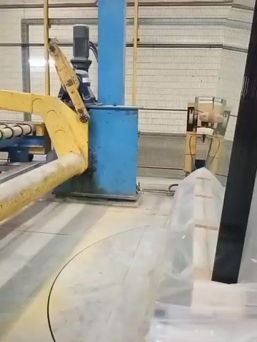 This video shows an industrial setting, likely a manufacturing plant in Spas-Zaulok, Russia. The primary focus is on automated machinery processing glass. A large, yellow, articulated arm is visible, extending from the left side of the frame. This arm appears to be a robotic manipulator or part of a conveyor system. It moves over a circular track on the floor, suggesting a rotational or carousel-like operation.

The machine it is attached to has several vertical yellow pipes, possibly part of a suction or lifting mechanism for handling large glass sheets. These pipes are equipped with metal clamps or rollers. In the background, a blue industrial unit with a motor on top is present, along with other yellow machinery and metal fencing that lines the walls of the facility.

To the right of the frame, stacked sheets of glass, partially covered in protective plastic, are visible. The lighting suggests an indoor environment, possibly during daytime hours. The overall impression is one of an active, automated industrial process. No people are visible in the video. No text is discernible in the frame.