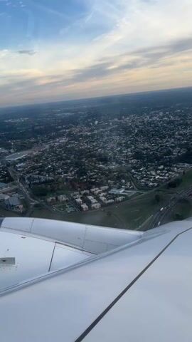 An airplane descends over a city, visible through the window and partially obstructed by the wing. The city is densely populated with buildings and trees. A network of roads and highways runs through the urban landscape. The sky is blue with scattered clouds, and the horizon is visible in the distance. The airplane wing is white and reflects the sunlight. The video is shot from inside the airplane, looking out the window.
