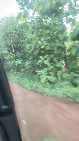 The video shows a view from inside a vehicle traveling on a dirt road. Lush green trees and foliage line the side of the road. The sky is visible with clouds. The camera appears to be mounted to the side of the vehicle, looking outwards. The movement of the vehicle suggests it is in motion.