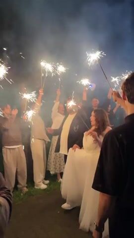 A group of people are gathered outdoors at night, holding up sparklers that are lit and producing bright sparks and smoke. In the center of the group, a couple is visible. The woman is wearing a white off-the-shoulder dress, and the man is wearing a dark suit jacket over a white shirt and scarf. The surrounding people are dressed casually, in various neutral-toned clothing. The background consists of dark trees and foliage, indicating an outdoor setting. The overall atmosphere appears festive, with the sparklers creating a celebratory ambiance around the central couple. The camera is positioned behind some of the attendees, looking towards the couple and the other guests.