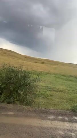 The video captures a dramatic skyscape over a rolling landscape in Karagayl, Kazakhstan. The scene is dominated by a massive, dark storm cloud that hangs low in the sky, with shafts of light piercing through to illuminate the distant hills. The terrain below consists of gently sloping, grassy hills, transitioning from vibrant green in the foreground to a more muted, dry yellow further up the slope. A dirt road, wet and reflecting puddles from recent rain, runs along the bottom of the frame, indicating a recent downpour or the approach of a storm.

The activity depicted is the dramatic passage of a weather system, with a powerful storm cell visible. The time of day appears to be late afternoon or early evening, given the low angle of the light and the atmospheric conditions. The weather is clearly unsettled and severe, with the dark clouds suggesting heavy rain, thunder, and possibly hail. The location cues—the vast, open grasslands and the style of the distant horizon with its darker, rugged formations—are consistent with the steppe environment of Kazakhstan.

There is no visible text in the video. The overall mood is one of awe and perhaps a touch of apprehension, evoked by the sheer power of nature displayed in the turbulent sky. The contrast between the dark, ominous clouds and the illuminated, rain-soaked landscape creates a visually striking and powerful image.