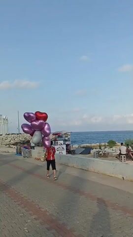 This is a vertical video compilation of scenes along a waterfront promenade in Yenişehir, Turkey.

The first clip features a young person in a red shirt and black shorts walking briskly while holding a large bunch of heart-shaped balloons in shades of pink, red, and silver. The promenade is paved with brick and bordered by a low concrete wall. Beyond the wall is the sea and a rocky shoreline. In the background, a person is seen near a kiosk or stall. The sky is blue with a few scattered clouds, suggesting a pleasant day.

The second clip transitions to a wider view of the promenade. An arched white structure, reminiscent of classical architecture, stands prominently on the left. The sea is to the right, with a rocky embankment separating it from the promenade. Several people are visible, some near stalls selling what appear to be souvenirs or merchandise. The sky remains blue with clouds.

The third clip shifts to a different section of the promenade, showcasing a dedicated blue bike lane with white markings. A person is riding an electric scooter on the bike lane, while a woman in a patterned dress walks on the adjacent paved pathway. Palm trees are visible on the left side of the frame, with the white arched structure from the previous clip visible in the distance. The time of day appears to be late afternoon or early evening, given the warm light.

Overall, the video captures the relaxed atmosphere of a seaside promenade, likely a popular spot for locals and tourists to stroll, cycle, and enjoy the coastal scenery. The presence of stalls and recreational activities suggests a lively public space.