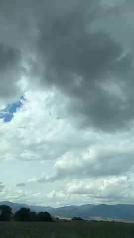 The video captures a wide shot of a cloudy sky above a rural landscape. The clouds are thick and gray, with some patches of blue sky visible. In the distance, rolling hills and mountains are silhouetted against the sky. The foreground is a field of dry grass. The video seems to be taken from a moving vehicle, as evidenced by the slight shake and the nature of the landscape passing by.