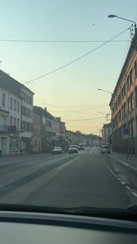 The video shows a view from inside a vehicle traveling down a street. The street is lined with buildings on both sides, appearing to be shops and residential buildings. There are several cars on the road, moving in the same direction as the vehicle. Traffic lights and street lamps are visible, as are power lines overhead. The sky is clear with a pale yellow hue, suggesting it is either sunrise or sunset. There are pedestrian crossings marked on the road. No people are clearly visible outside of the vehicles. The overall scene depicts a typical street in a town or small city.