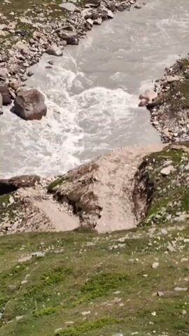 A drone-like aerial view pans across a wide, turbulent river flowing through a mountainous, rocky, and grassy landscape. The river appears muddy and is flowing rapidly, with visible rapids and churning water. The banks of the river are steep and eroded in places, with loose rocks and soil. Patches of green grass and vegetation are present on the slopes, interspersed with large rocks and boulders. The terrain suggests a remote, natural environment. The video begins with a close-up of the river and its banks before pulling back to reveal the broader valley and surrounding mountains under a partly cloudy sky. There are no people or man-made structures clearly visible in the scene.