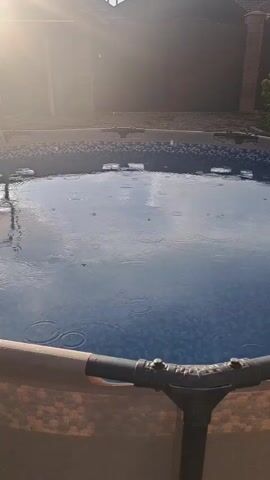 The video shows an overhead view of an above-ground swimming pool during a rain shower. The sun is shining brightly, creating a glare on the water and casting shadows. Raindrops are visible hitting the surface of the pool, creating ripples. The edge of the pool, made of a brown material and supported by metal pipes, is in the foreground. In the background, there are some buildings, trees, and power lines. There is also a pool net hanging on a pole over the water. The overall scene is peaceful, with the sound of rain and the gentle ripples on the water.