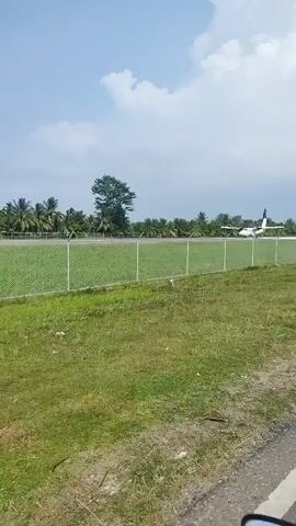 A white twin-engine turboprop aircraft with orange accents is taxiing on an airport runway, viewed from the side of a road separated by a green-fenced area. Palm trees and other foliage are visible in the background under a blue sky with scattered clouds. The camera is positioned to the right of the fence, looking towards the runway.