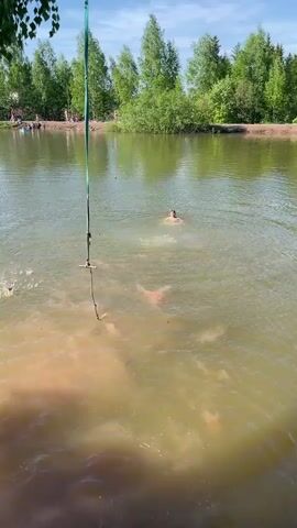 A vertical video shows a young man swimming in a murky pond. The water is green and brown, with some ripples. He is shirtless, with only his head and shoulders visible above the water. In the foreground, a rope swing hangs from a tree, with a wooden seat at the end. In the background, there are trees and other people on the shore. The sky is blue with some clouds.