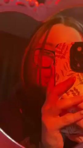 The video shows a person taking a selfie in a heart-shaped mirror. The room is lit with red light, creating a red tint on the person and the phone. The person is wearing glasses and holding a phone with a floral patterned case. The person's face is partially obscured by the phone. The mirror has an ornate edge, illuminated with a soft red glow. The person seems to be standing still while holding the phone to take the photo. There is background noise, including someone speaking in a foreign language.