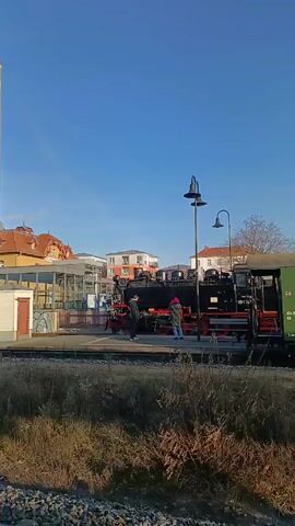 The video shows a black steam locomotive, numbered 89 7467-7, at a train station on a sunny day. The locomotive has red wheels and some steam is coming from the top. Two people are standing near the locomotive on the platform. There's a green passenger car attached behind the locomotive. The station has a sheltered waiting area and older buildings with orange roofs in the background. Overhead power lines are visible above the tracks. The foreground has weeds and gravel.