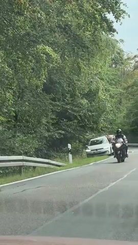 A dark colored motorcycle with a rider wearing a black helmet and black and white riding suit is traveling on a paved road. The road is lined with a metal guardrail on the left side and a dense green forest on both sides. A white car is visible in the distance on the right side of the road, also traveling in the same direction. The camera appears to be positioned inside a vehicle, looking out at the motorcycle.