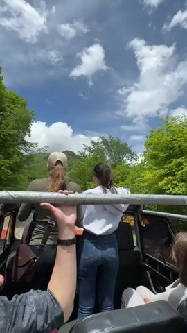 The video shows a group of people in the back of an open-air vehicle, likely an SUV or jeep, driving on a dirt road. The perspective is from within the vehicle, looking out. Two young women are visible in the foreground, standing and holding onto a metal bar for stability. The woman on the left has long blonde hair and wears a tan baseball cap and a green t-shirt. The woman on the right has dark hair in a ponytail and wears a grey hoodie and blue jeans. Another person is partially visible in the lower right corner, wearing a light-colored shirt. The vehicle is moving through a lush, green environment with trees lining the road and mountains in the background. The sky is blue with scattered white clouds. The road is rough and appears to be made of gravel and dirt. The video captures the movement of the vehicle and the surrounding scenery, suggesting a journey through a natural landscape.