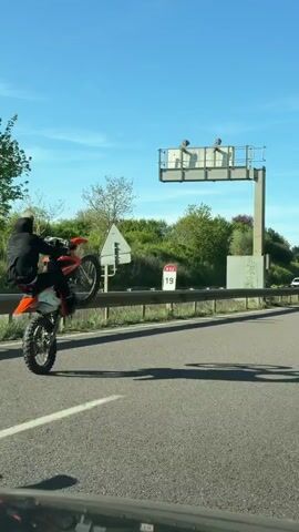 A video shows three motorcyclists riding on a multi-lane highway on a sunny day.  The motorcyclist in the lead is riding an orange dirt bike and performing a wheelie. The second motorcyclist is riding a blue and white Yamaha dirt bike, while the third is riding a green dirt bike. They are all wearing helmets and casual clothes. There are trees, a guardrail, and a highway sign to the side of the road.  A white car can be seen in the distance. The video is taken from inside a car that is driving alongside the motorcyclists.