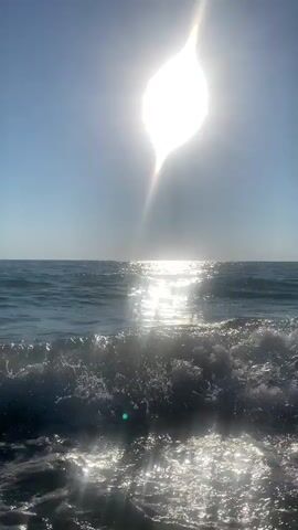 The video displays a close-up, low-angle perspective of ocean waves crashing near the shore. The sun is prominently positioned in the upper center of the frame, emitting a bright, lens-flared glow. The sky is clear and blue. The water is a deep blue, and the sun's reflection creates a shimmering path across the surface. The foreground is dominated by white foam and spray from the breaking waves, suggesting movement and the dynamic nature of the sea. There are no people or man-made objects visible in the scene. The lighting indicates it is daytime, likely mid-morning or afternoon, given the sun's high position.