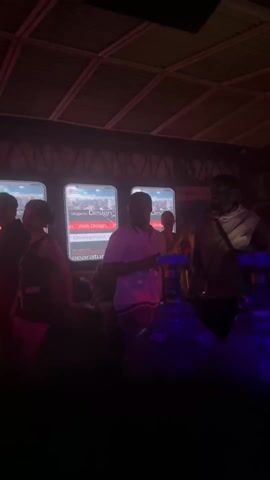 The scene is indoors in a dimly lit, crowded space that appears to be a nightclub or bar. There are several people visible, though many are obscured by the low light. The primary lighting consists of blue and red lights. In the foreground, several large, cylindrical containers are visible, seemingly tanks. In the background, there are displays with the words "Skypros Design - Web" and what looks like "Develop". People are moving around and socializing, with one person wearing a light-colored hoodie and a crossbody bag being particularly noticeable. The structure above looks like it may be corrugated metal.