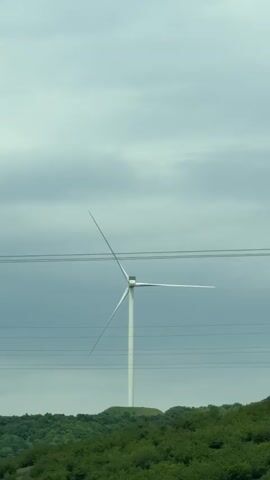 The camera, likely inside a moving vehicle, films a wind turbine against a gray, overcast sky. A lush, green landscape is visible at the base of the turbine. Power lines intersect the view, adding a layered visual element. As the vehicle moves, a speed limit sign reading "110" briefly enters the frame. The overall impression is one of traveling through a rural area on a cloudy day, where renewable energy is being generated.