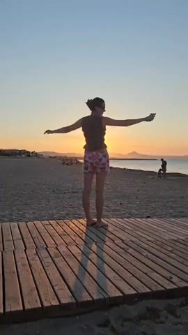 A person stands on a wooden walkway at the beach facing away from the camera, towards the ocean and a sunset. The person has their arms outstretched. The sky is clear and blue with a bright orange sunset on the horizon. There are a few other people on the beach in the distance.