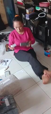 A woman with her hair in a bun and wearing a pink long-sleeved shirt and black pants sits on a tiled floor surrounded by packages. She holds a small item in her hands and appears to be examining it or preparing it for packaging. A roll of tape, scissors, and a cell phone are visible on the floor near her. In the background, a dark cabinet with items on top and a glimpse of clothing are visible. Another person's bare foot is visible in the lower right corner of the frame. The woman is engaged in what appears to be the process of packing or preparing items for shipment.