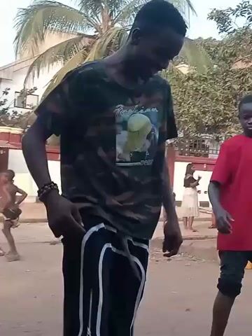 The video shows a scene outdoors with two young men, one wearing a red t-shirt and the other a camouflage shirt, facing each other and kicking a soccer ball back and forth. Several children are also present, some watching and others playing near a table with items on it. Banana trees and other foliage are visible in the background. The ground appears to be dirt or sand.