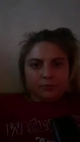The video shows a close-up of a young woman's face. She is wearing a red shirt with white lettering. The lighting is dim and reddish, casting shadows on her face. Her hair is messy and pulled back. She makes several facial expressions, including a neutral gaze, sticking out her tongue, puckering her lips, and a slight smile. In the last frame, she turns her head slightly to the side, and her eyes appear to be closed or looking down. A dark object, possibly a phone or remote, is visible in the lower right corner of the frame. The background is a plain, light-colored wall.