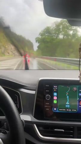 The video is taken from inside a car driving on a rainy day. We see the dashboard, steering wheel, and windshield wipers in action, clearing the rain. A navigation system is visible on the console indicating the route. Outside, other cars are visible on the wet road, and the scenery is dominated by a forested hillside. A cross hanging from the rearview mirror is visible at times.