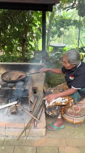 Elderly woman roasts coffee beans over traditional oven in Tampaksiring