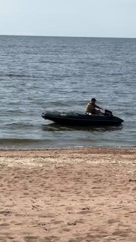 A person is in an inflatable boat with a motor on a body of water. The boat is moving, and the person is operating the motor. The boat is near a sandy beach.
