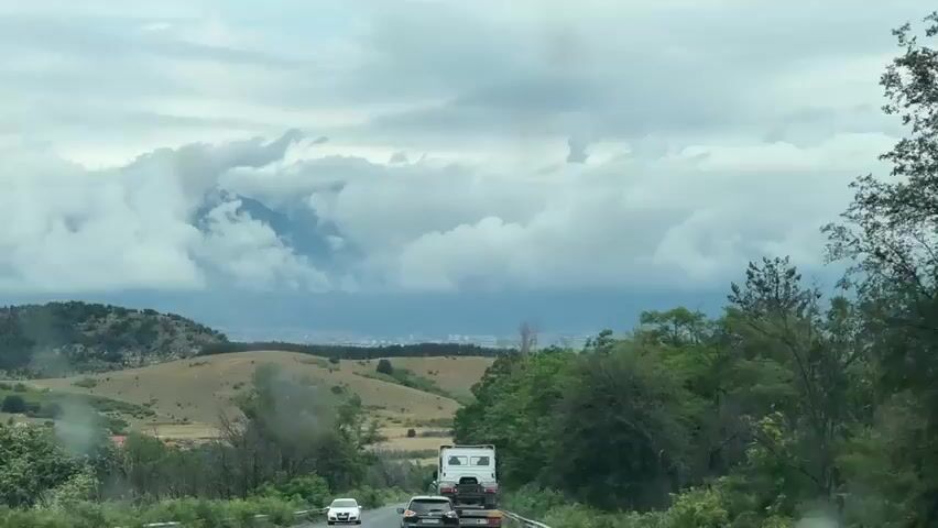 A vehicle's dash cam footage shows a cloudy sky over a rural landscape with hills and trees. A road with other vehicles, including a white truck with a camper shell and a few cars, is visible. In the distance, a town or city can be seen. The clouds are heavy and appear to be covering a mountain range in the background. The footage appears to be taken during daylight hours.