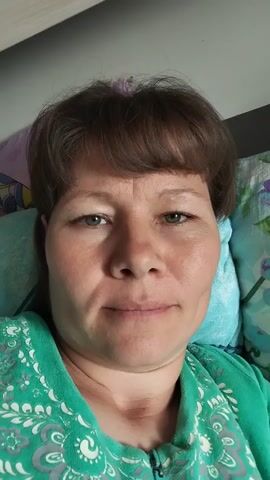 A woman with brown hair and bangs lies on her side in bed. She is wearing a green shirt with a white floral pattern. Her face is the focus of the video. The video captures her making various facial expressions, including winking, smiling, and widening her eyes. The lighting is natural, coming from an unseen source. The background shows bedding.