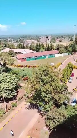 The video shows an aerial view of a landscape, likely a campus or school environment, with buildings, trees, and a clear blue sky.  The camera pans to the left, revealing more of the landscape, including a small, round structure in the distance and more buildings interspersed among the trees.  A road with two people walking on it is briefly visible at the bottom of the frame.