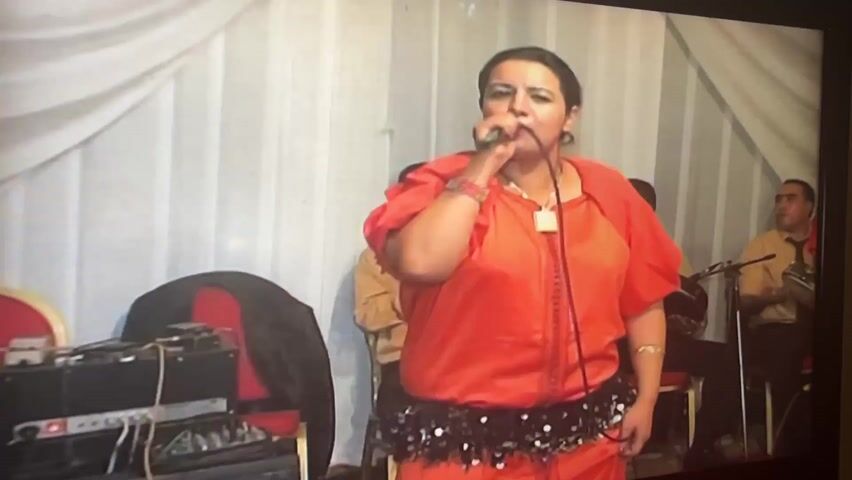 A woman in a bright orange tunic and black sequined belt is performing into a microphone, with a band behind her. She has dark hair pulled back and is wearing jewelry. The band consists of at least three men playing instruments, one on a stringed instrument and another on what appears to be a keyboard. The background features white drapery and the scene is cluttered with musical equipment. The video appears to be recorded from a screen.